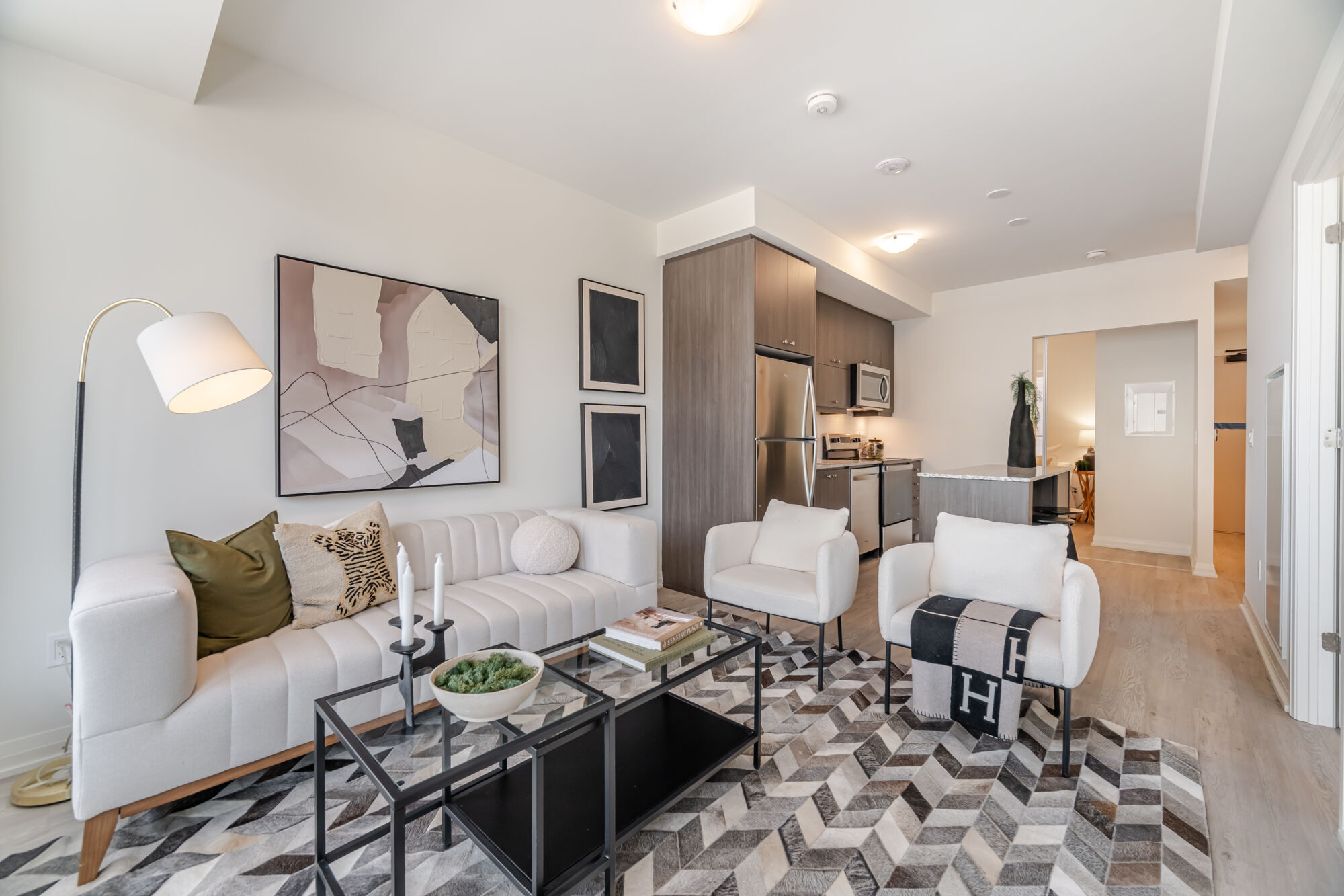 Condo living room with a white couch, 2 white side chairs, 2 glass nesting tables and a dark herringbone rug. Kitchen with dark wood cabinets, kitchen island, quartz countertop and stainless steel appliances. 
