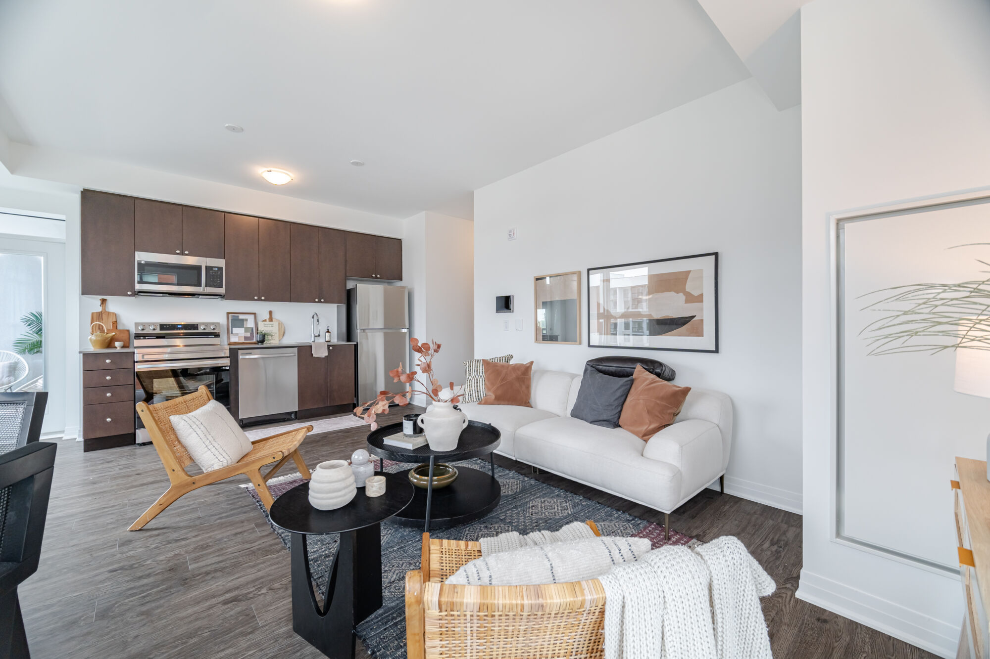 Condo Kitchen and and living room. Kitchen features dark cabinetry and stainless steel appliances, living room features white fabric couch, wicker chairs black accent tables and a patterned rug. 