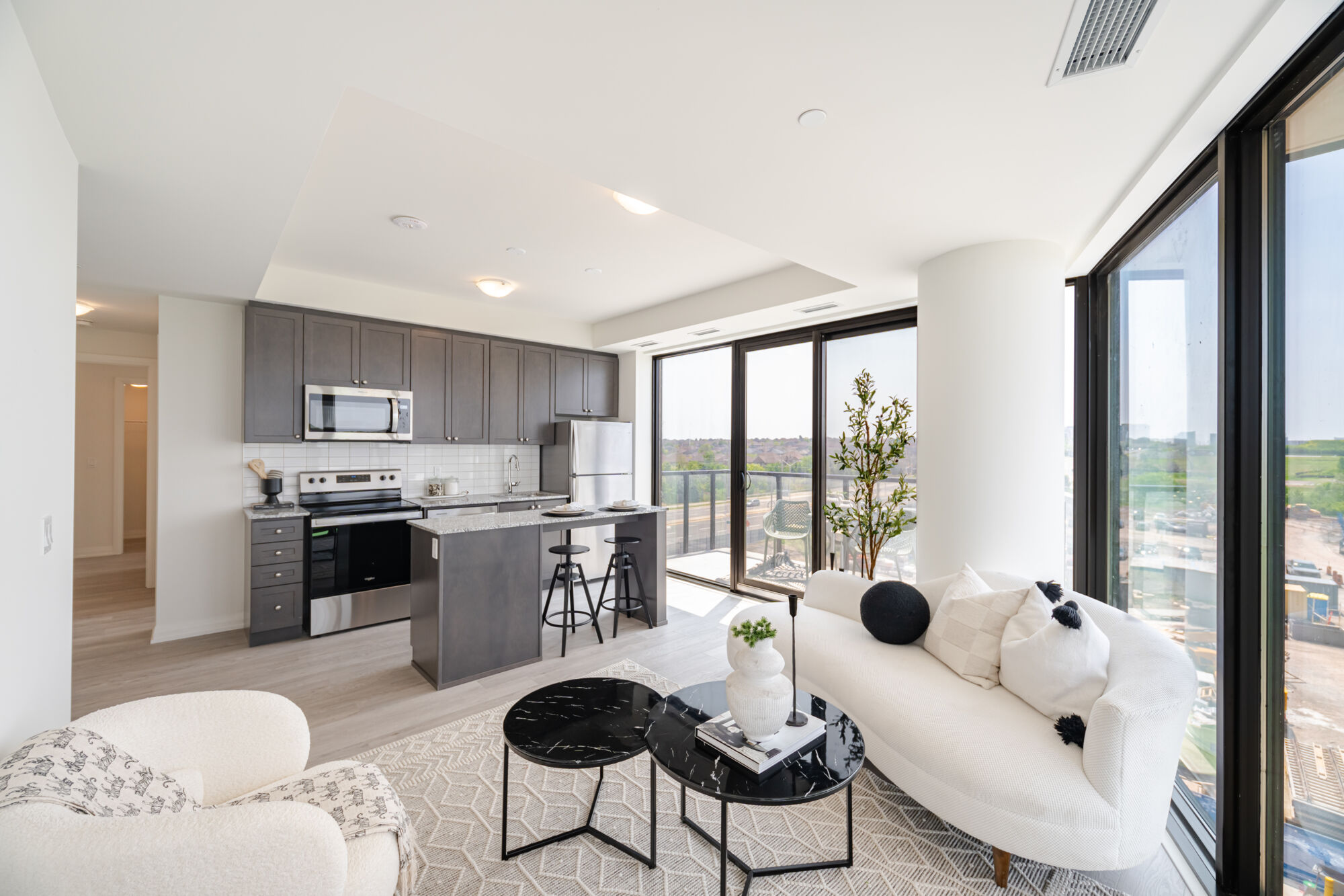 Condo kitchen with dark cabinetry, stainless steel appliances, kitchen island and granite countertops. Living room with white couch and carpet, black coffee tables and pillows. 
