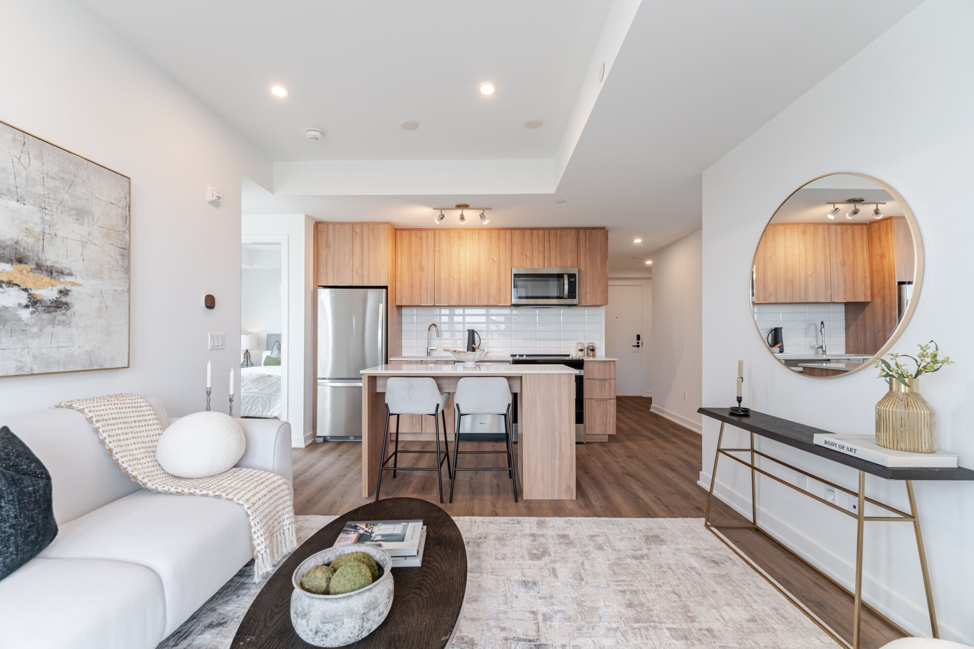 Condo Kitchen with wood-grain cabinetry, stainless steel appliances, kitchen island and quartz countertop. Living Room with a white couch, decorative pillows and blanket, neutral rug and dark wood coffee table. 