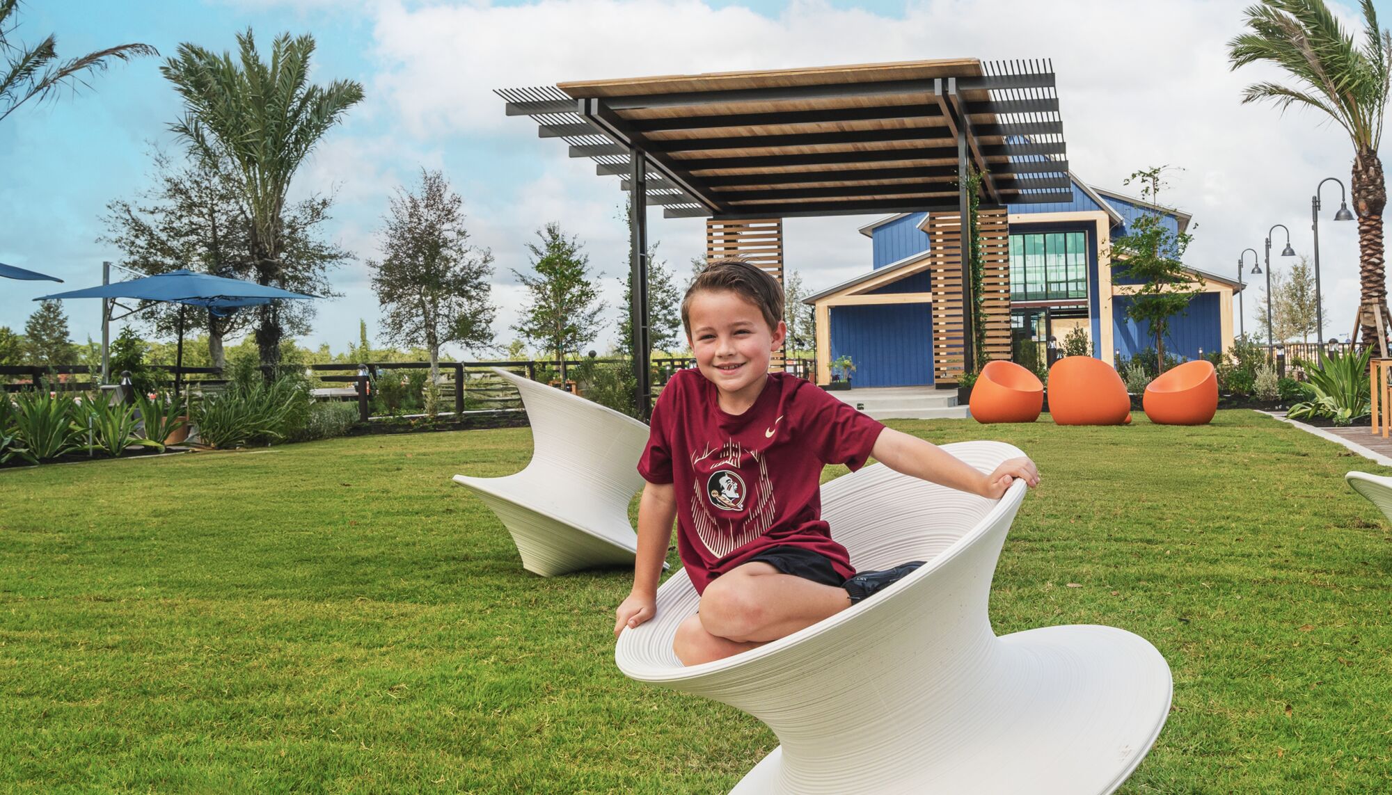 Child sits on a spinning top-style chair in a community yard with bright green grass, surrounded by palm trees.