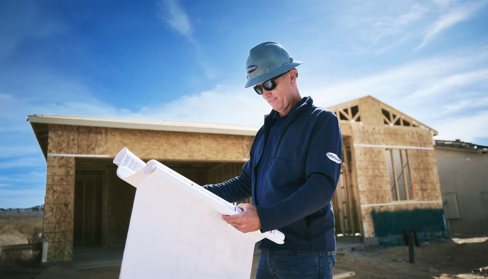A builder wearing Mattamy clothing stands in front of a home under construction looking at blueprints.