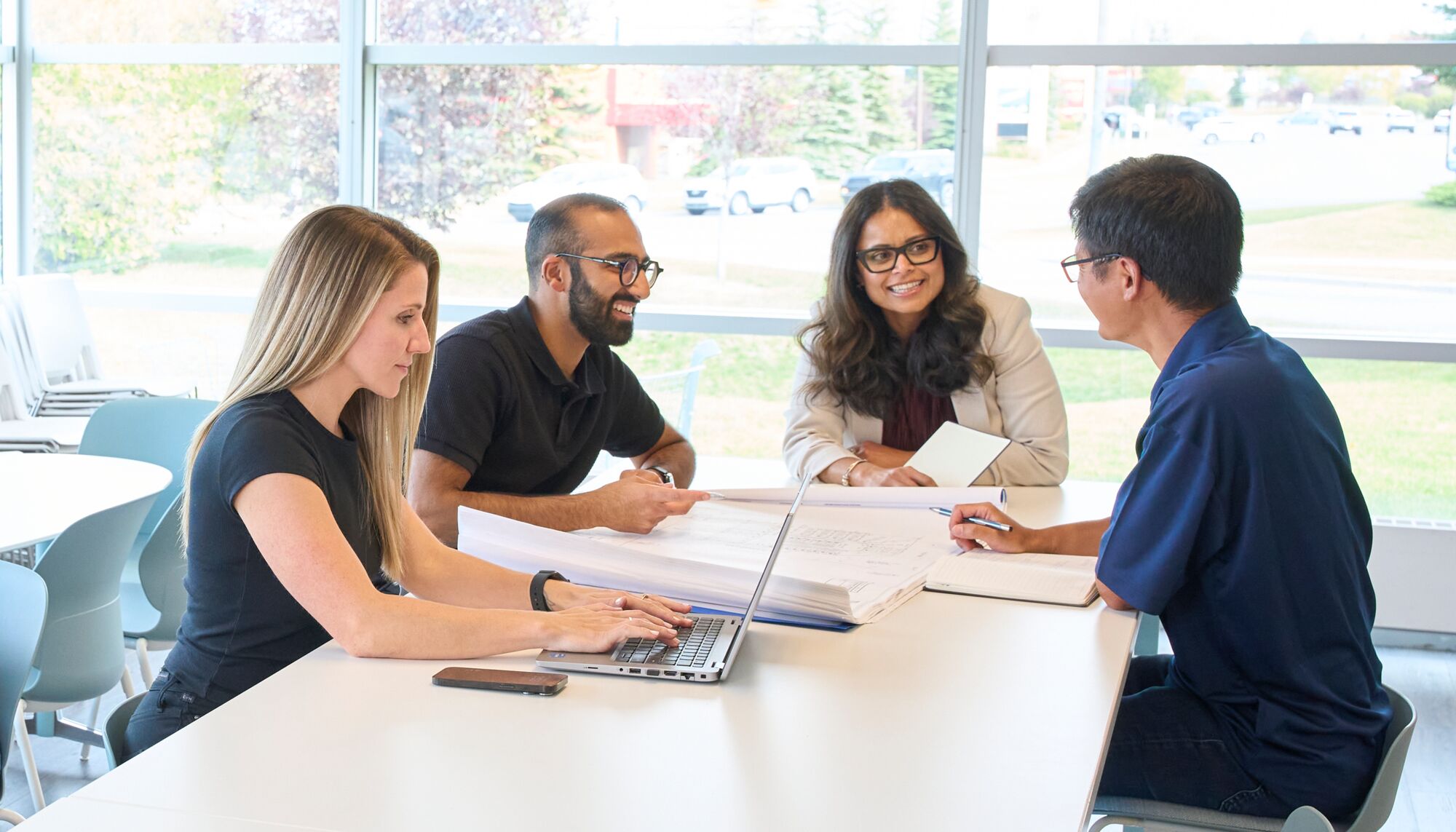 Four office workers, two men and two women sit at a table infant of a laptop and various blueprints smiling and working together.