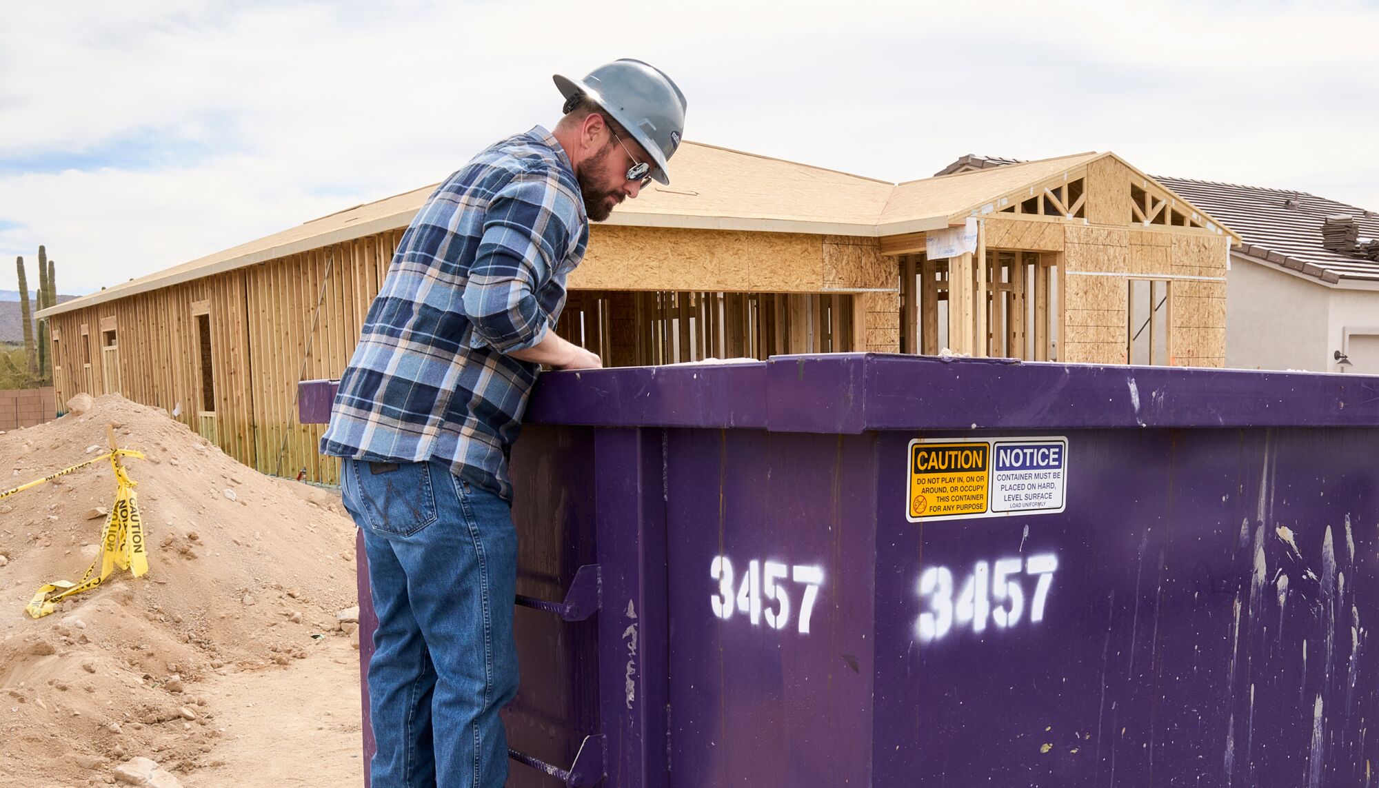A man stands over a drywall recycling dumpster in front of a framed home