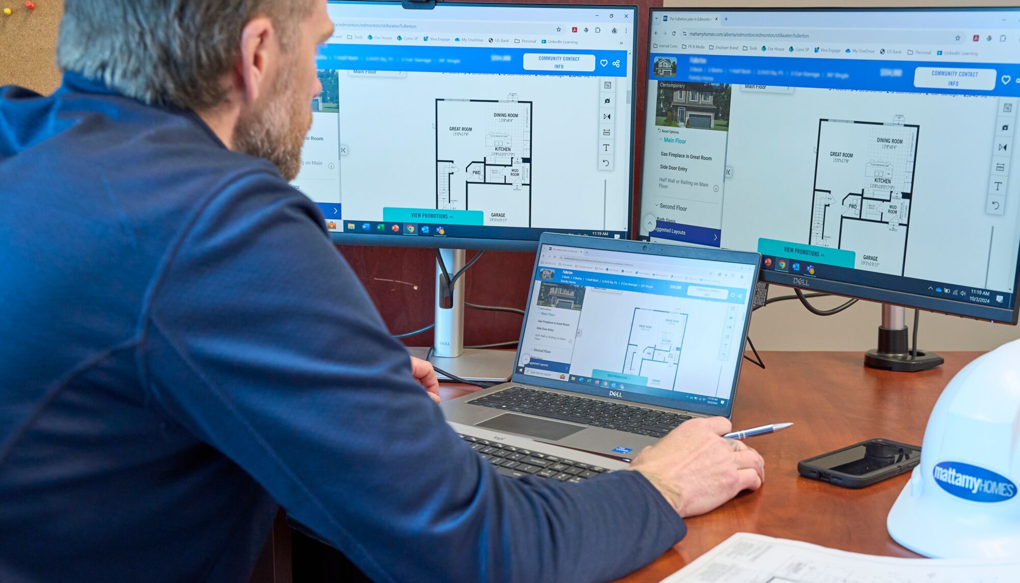 A man sits at a desk with a laptop and two monitors looking at home floorplans