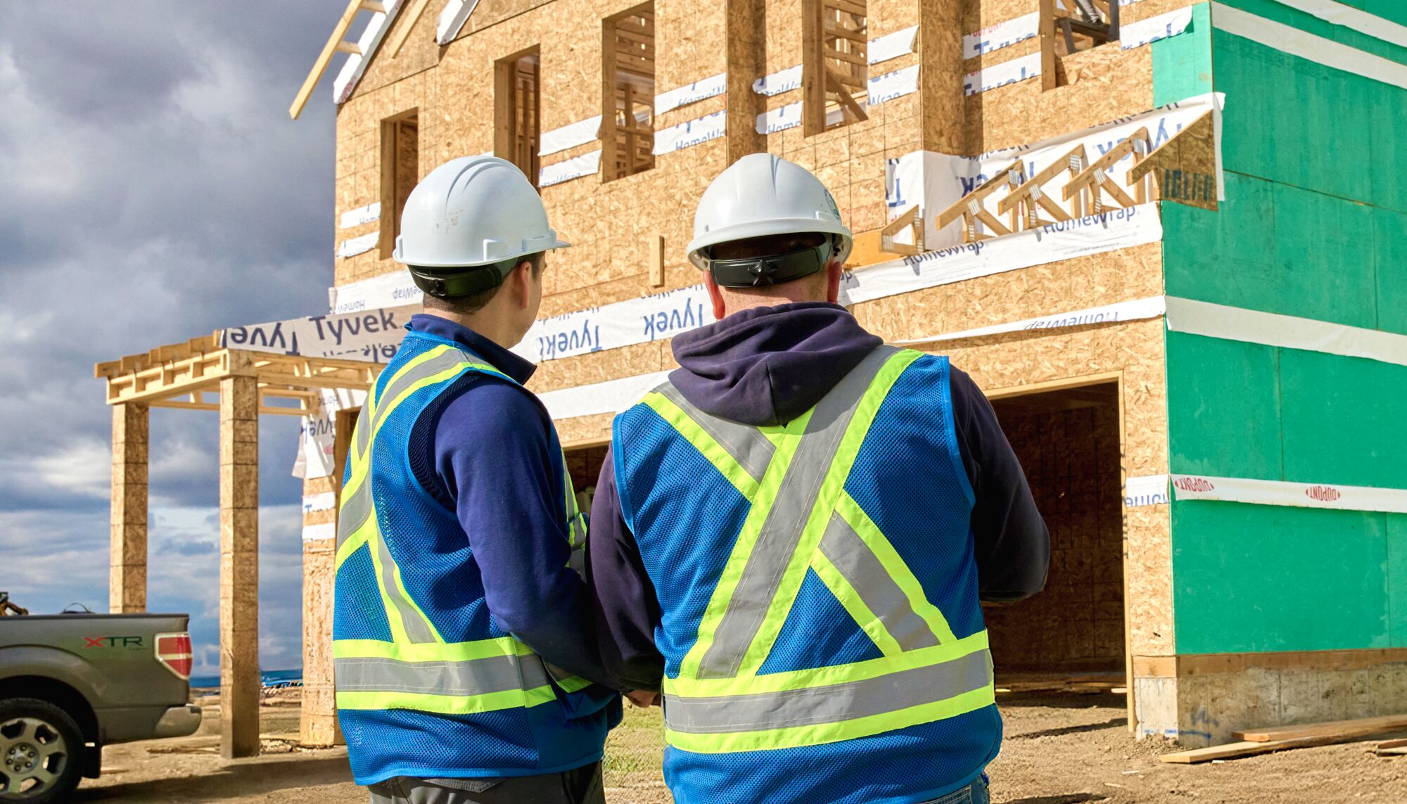 Two workers stand in safety vests and hard hats in front of a framed home