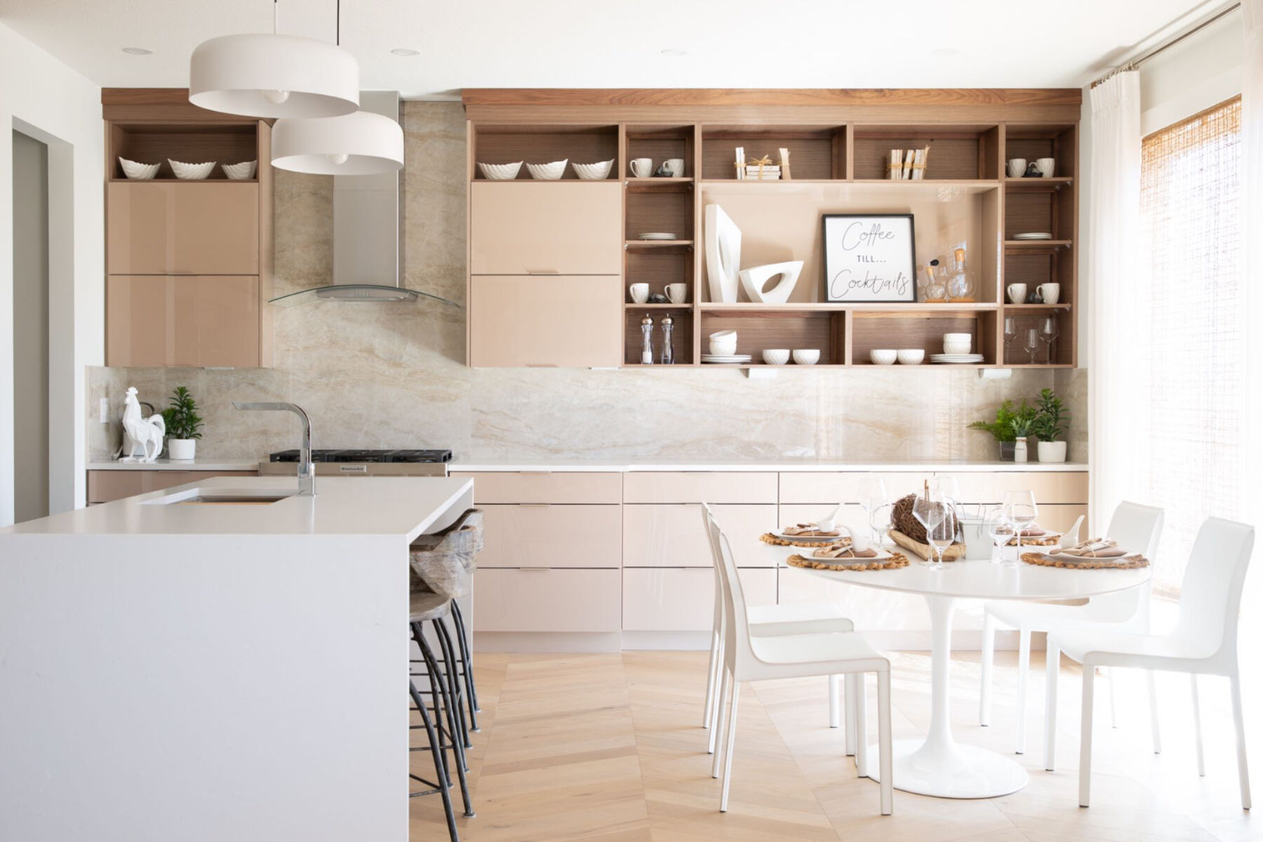 Kitchen and dining area with light walls, wood chevron floors, pendant light above island, light beige tile backsplash, steel appliances, patio window on right, beige cabinets and drawers, and a white island and dining set