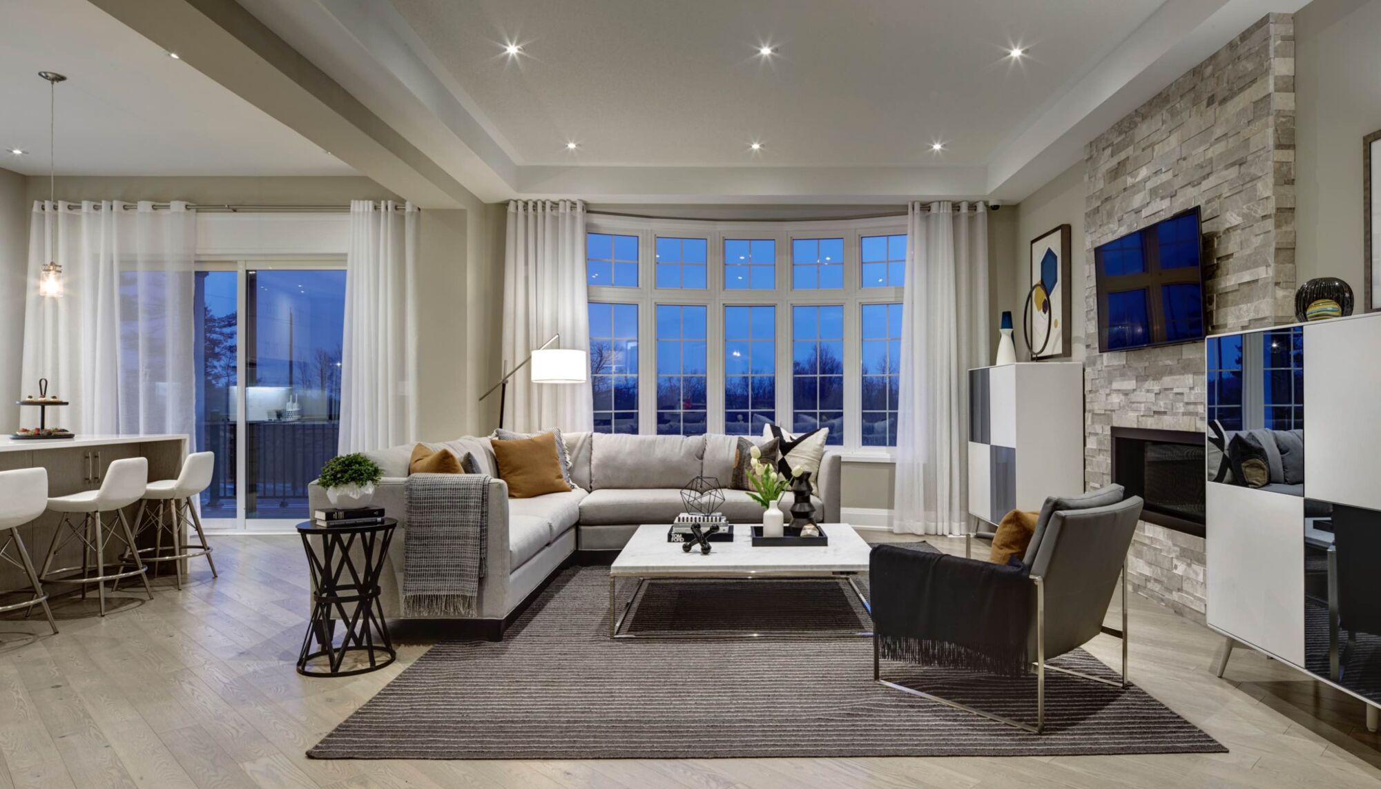 Living room with a grey L-shaped couch and armchair facing a television, against a large window.