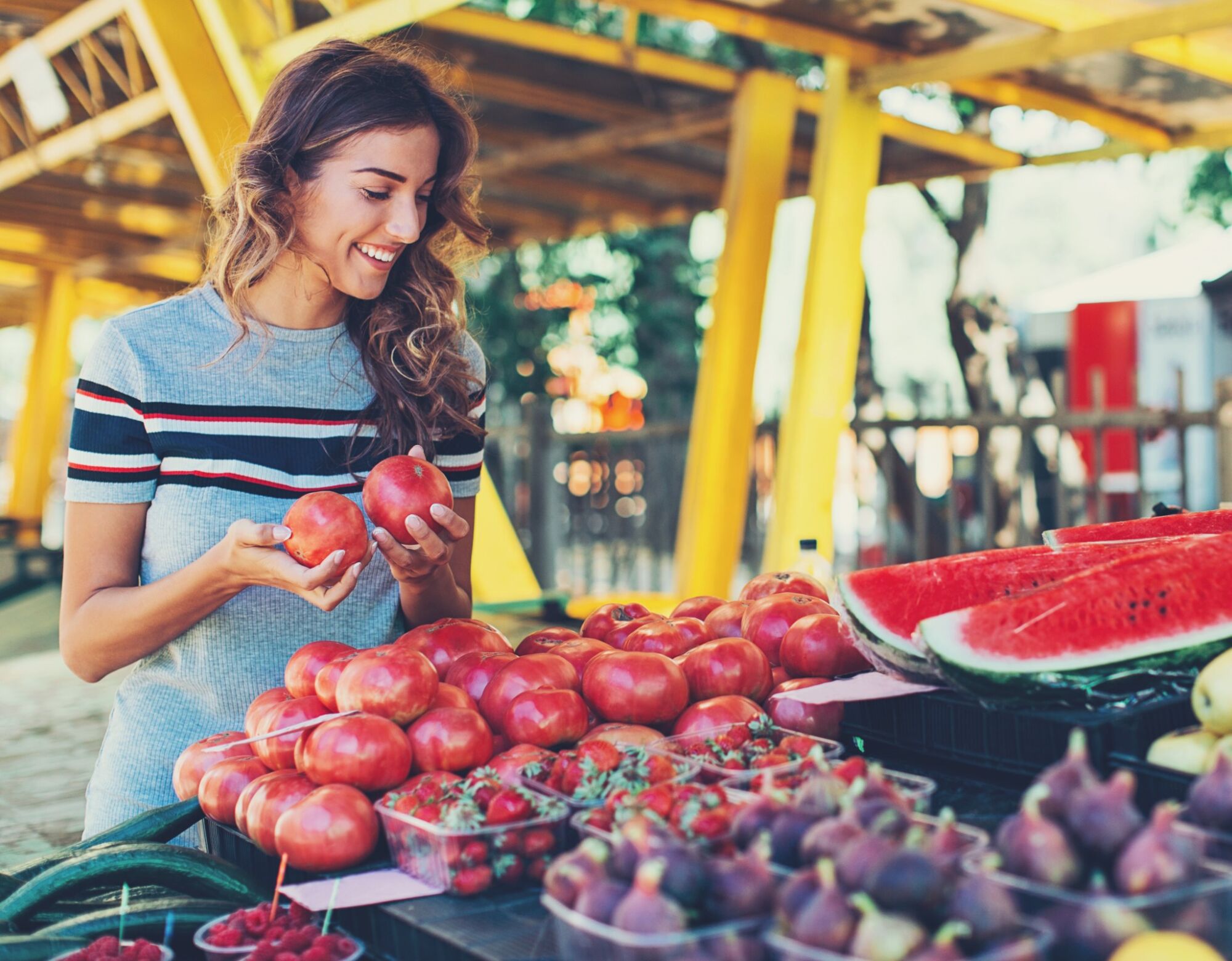 A woman picks out tomatoes while surrounded by fruits at a farmer's market.