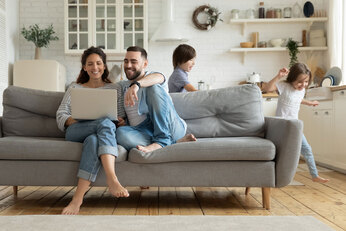 A man and woman sit on a couch while children run around in the background.