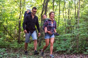 Family of four, with dad carrying baby on back in bag, and mom carrying older child on back, hiking in a forest surrounded by greenery