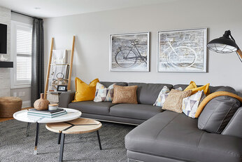 Living room with light walls, wood floors, light tile fireplace with mounted tv above, window beside fireplace with grey curtains, grey L-shaped couch with yellow and white throw pillows facing tv